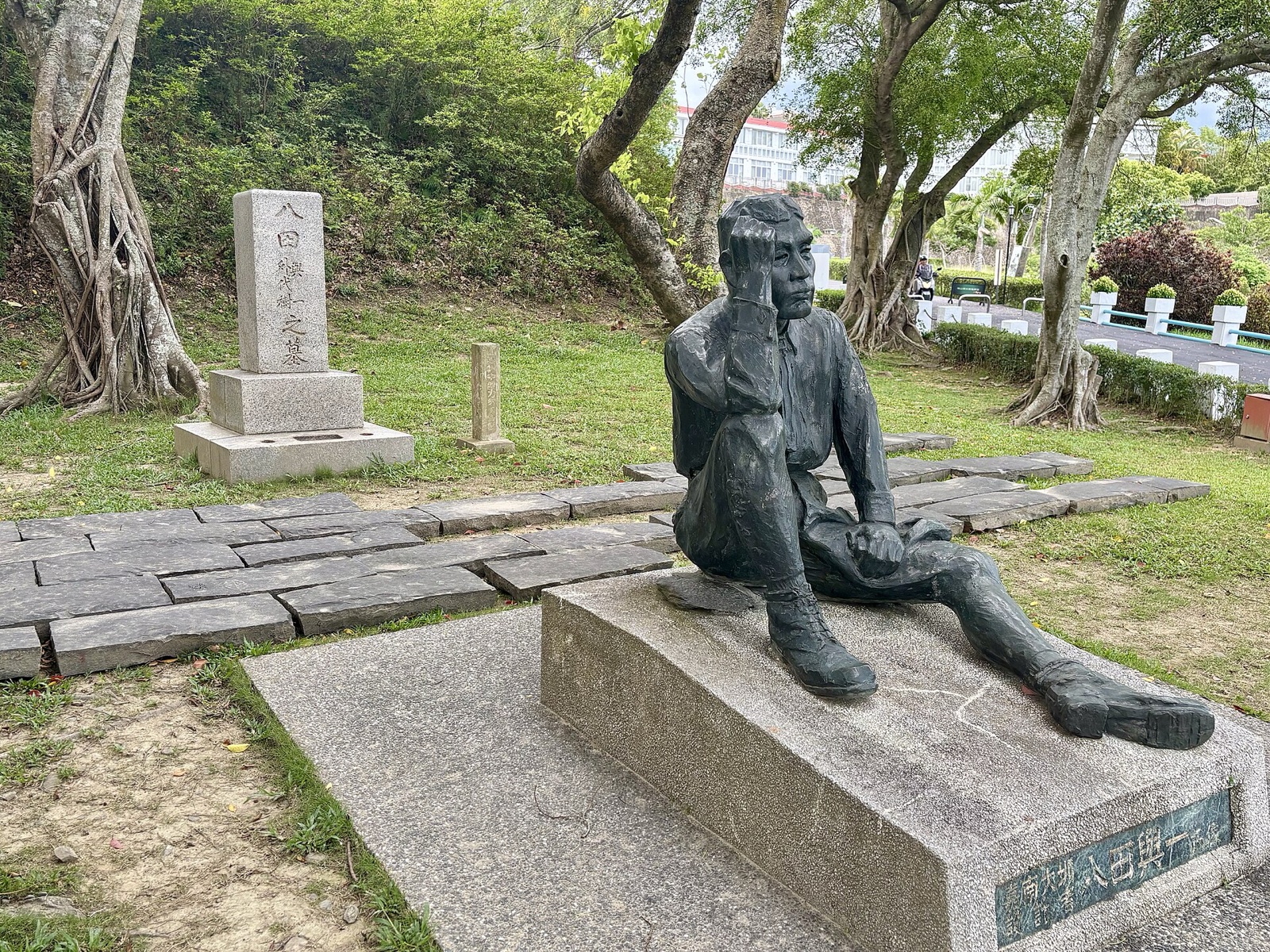 烏山頭の八田與一像。片手を頭に当てて座る銅像の後ろに、木立の中の石の墓碑が見える。写真：Mrmarkertw / Wikimedia Commons / CC BY 4.0（2025年6月1日撮影）