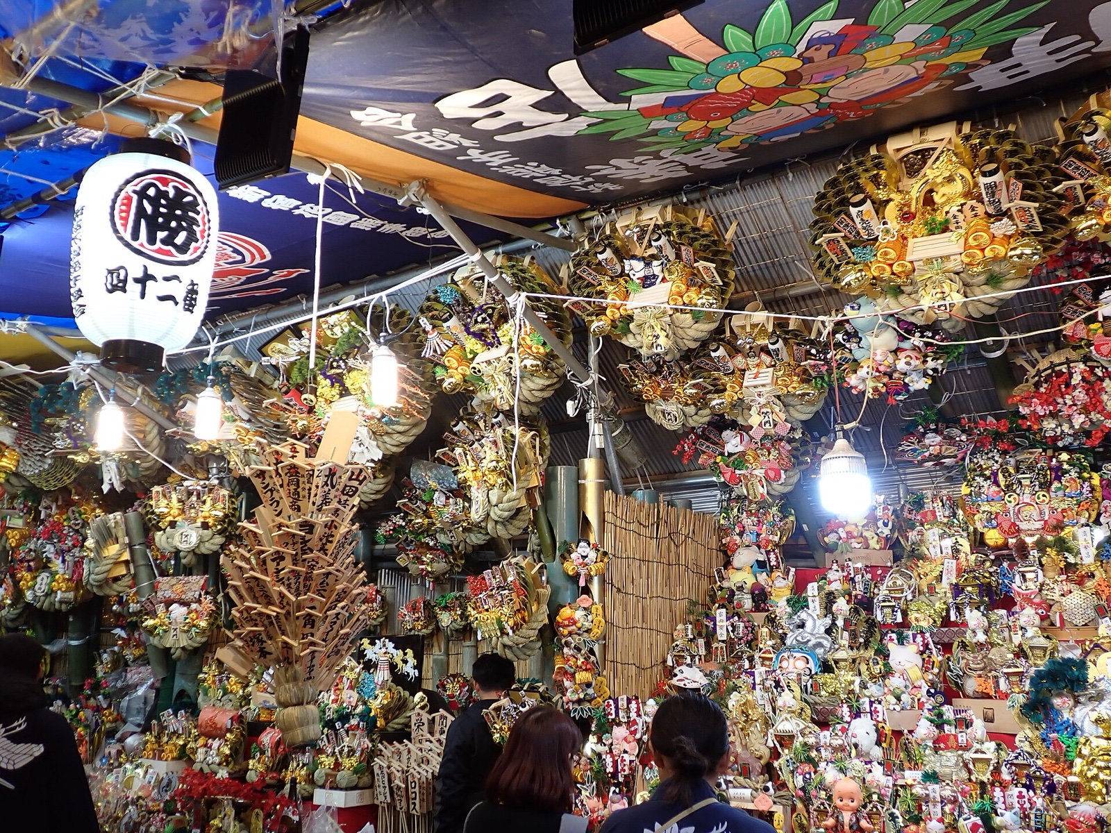 花園神社の酉の市の熊手店。「勝」の提灯の下に、おかめや宝船を飾った熊手が天井まで並ぶ。写真：Mozzucu / Wikimedia Commons / CC0（2025年11月24日撮影）