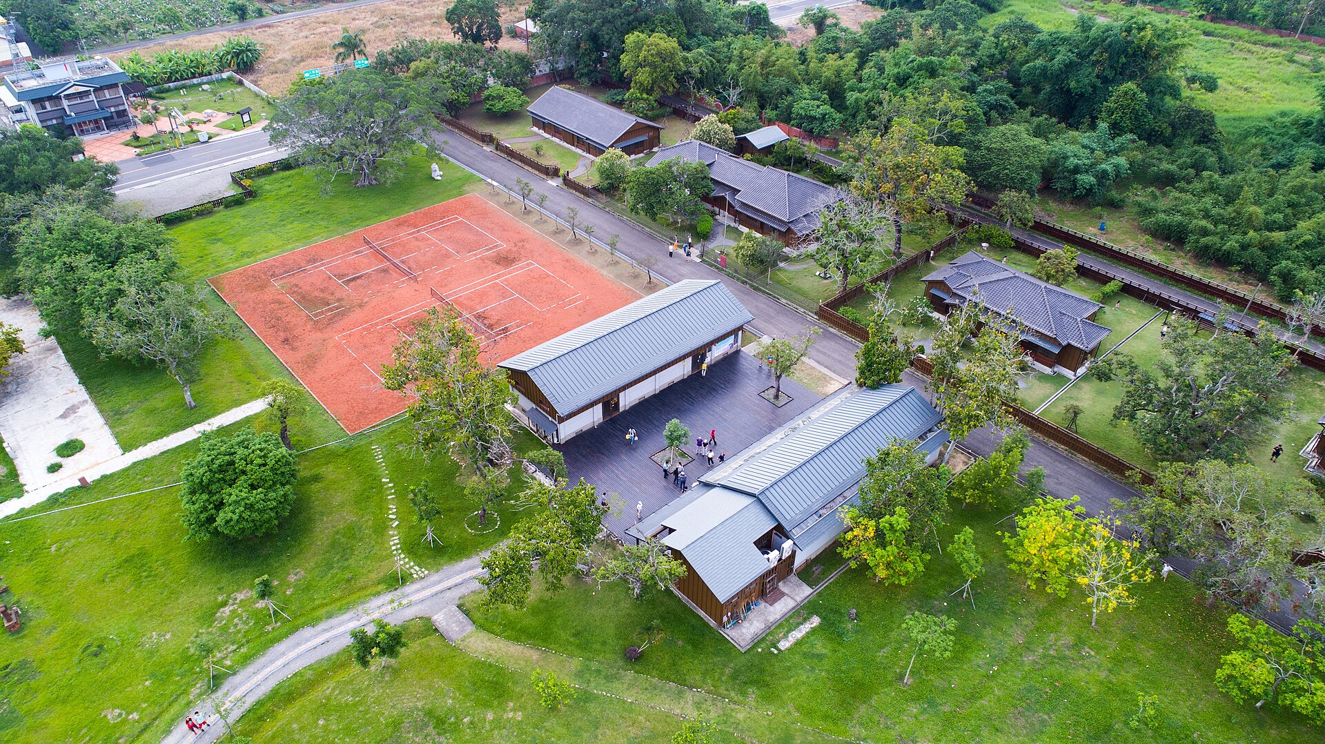 空から見た八田與一紀念園区。芝生と木立の中に灰色の屋根の日本家屋が並び、そばにクレーのテニスコートがある。写真：Guanting Chen / Wikimedia Commons / CC BY-SA 4.0（2017年5月28日撮影）